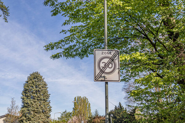 Traffic sign: 30 km zone ends, white sign with the number 30 inside a gray circle and three black lines crossing it, trees with lush green foliage and a blue sky in the background