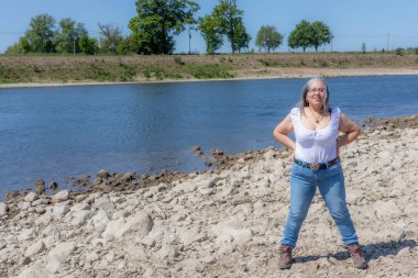 Gülümseyen olgun bayan yürüyüşçünün Maas Nehri kıyısında duraklaması, Maasvallei doğa rezervi, kot pantolon, gri saç, arka planda ağaçlar, Hollanda 'nın güney limanında güneşli bir gün. Etkin yaşam konsepti