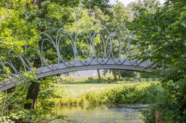 Nehir üzerindeki gri metal köprü yemyeşil ağaçlar ve vahşi bitki örtüsüyle çevrili yeşil çimenler bulanık arka planda güneş ışığıyla aydınlatılmış, Hollanda 'nın Güney Limburg kentinde güneşli bir bahar günü.
