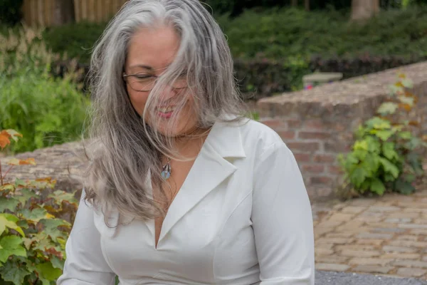 Portrait of mature woman with smiling expression, looking down, hair disheveled and covering face, straight gray hair, tanned skin, eyeglasses, light makeup, green vegetation in background