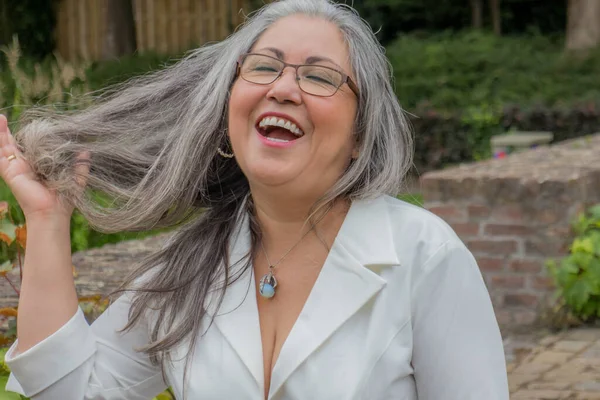 Portrait of mature woman with broad smile, eyes closed, head back, touching straight disheveled gray hair, tanned skin, glasses, light makeup, relaxed day with green vegetation in background