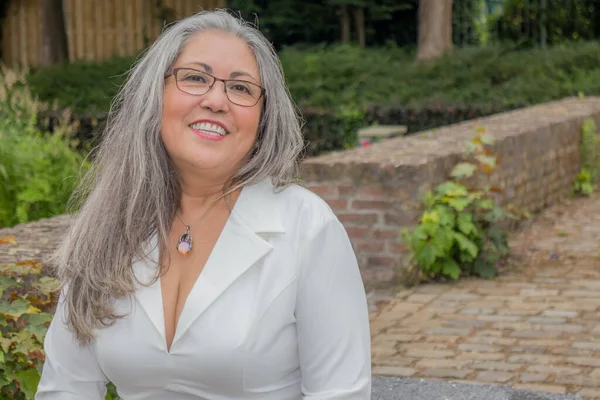 Portrait of mature woman with smiling expression, head turned slightly, attentively looking at camera, straight gray hair, tanned skin, glasses, light makeup, green vegetation in background