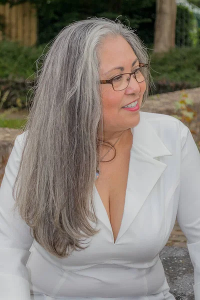 Portrait of a mature woman with a calm expression, slight smile, head turned in profile, looking attentively, straight gray hair, tanned skin, glasses, light makeup, green vegetation in the background