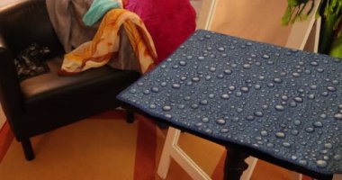 Adult female hands placing a straightener iron, black brush and brown comb on a small table with a blue surface, for straightening hair, room with a bench, a mirror and scattered multi-colored scarves