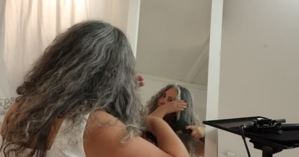 头发蓬松的成熟女人在镜子前看着自己 开始用刷子梳理她的头发 开始整理她那波浪形的灰黑色头发 白色背景的过程 — 图库视频影像