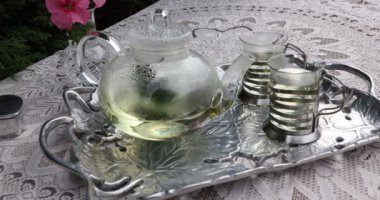 Metal tepside nane çayı, yeşil çayla çaydanlık, pembe çiçeklerle dolu bir vazo, ince kahverengi bir masa örtüsü taşıyan bir masa. Bahçede çay zamanı konsepti