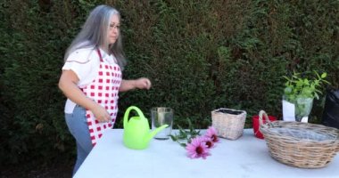 Mature woman working on a worktable in the garden of a house against a green bush in the background. Purple flowers, glass vase, wicker pots, green plastic watering can and plants on a white surface