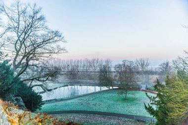 Winter landscape of the Dutch countryside against bare trees and a blue sunrise sky, seen from a hill at a lookout point, pond, frozen green grass, winter day in Kasteelpark, Elsloo South Limburg, Netherlands