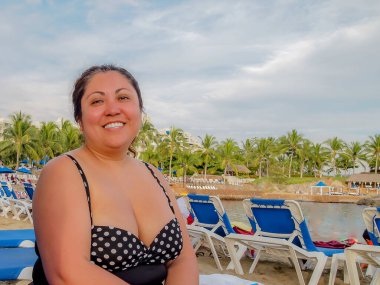 Orta yaşlı Meksikalı kadın portresi gülümsüyor, mayo giyiyor ve bulutlu gökyüzüne bakıyor, kameraya bakıyor, siyah bağlı saçlar, güneşlenen sandalyeler ve bulanık arka planda plajlar, tatilin tadını çıkarıyor.