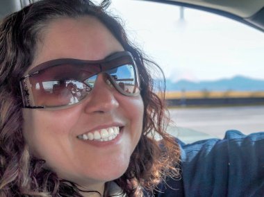 Middle-aged Mexican woman with the photographer reflected in the sunglasses, driving her car, looking at the camera with a blurred background, wide smile, brown hair, sunny day on the road in Mexico
