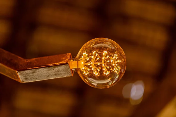 Antique decorative lamp lit against soft blurred yellowish golden brown background, filaments showing through glass, steel lamp holder on ceiling. Led technology