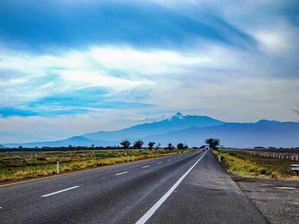 Mexican road landscape against Colima volcano with a cloudy sky, straight asphalt road with white line markings, green valley in the background, summer day in Jalisco state, Mexico