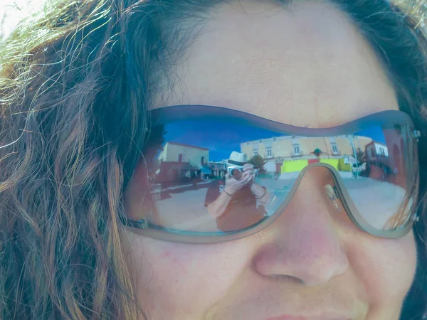 Close-up scene of a woman\'s face wearing sunglasses reflecting the photographer taking the photo, buildings in the background against a blue sky, middle-aged woman, wavy brown hair, man wearing a hat