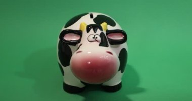 Front view of Adult female hand placing coins into a piggy bank shaped like a cow against a green  studio background. Concept of saving for the future, dreams, emergencies and prosperity