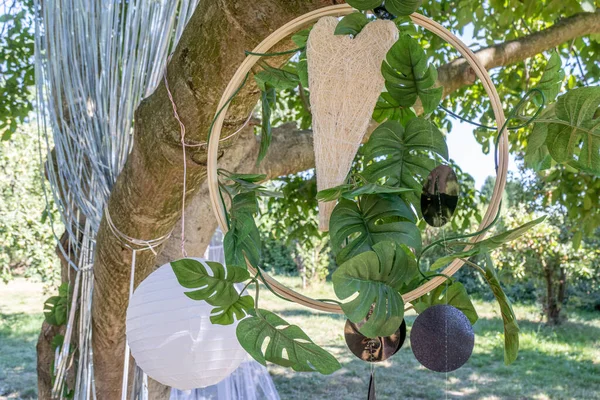 A straw heart in a wooden hoop, hanging on a tree against lush green foliage leaves, blurred background, white paper lantern, black circles, romantic decoration for a garden wedding