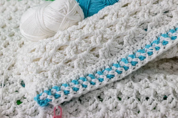 Edge of a white crochet blanket with unfinished blue detail, embossed crochet stitch pattern, pink stitch marker, one blue and one white cotton ball in the blurred background. Handmade creativity