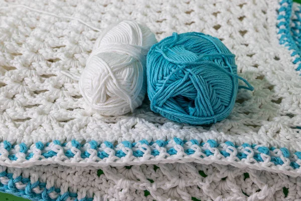 Front view of two skeins, one white and one blue, metallic pink crochet hook on white crochet blanket, blue details on edges, blurred background, raised crochet stitch pattern. Handmade creativity