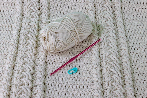 A cotton ball, pink metal hook and green stitch marker on a beige cotton yarn crochet blanket, interlaced braids, original raised crochet stitch pattern. Handmade craft creativity