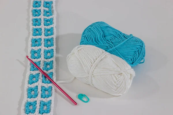 Crochet fabric interlacing the colors white and blue next to two cotton balls, a pink crochet hook and green stitch markers against a white background, pattern in square shapes. Handmade creativity