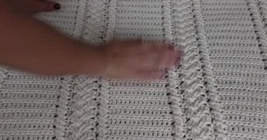 Surface of a crochet blanket with intertwined braids finished in beige cotton yarn, embossed stitch pattern, adult female hands touching it, red nails. Handmade craft creativity