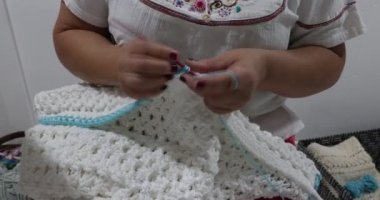 Elderly female hands knitting the border in blue cotton yarn on a white crochet blanket, pink metal hook, original embossed crochet stitch pattern. Handmade craft creativity