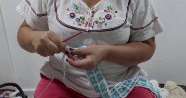 Female adult hands begin to crochet with the blue thread a blanket in the process of being made, simple pattern of interlocking squares in white and blue colors, pink metal hook. Handmade creativity