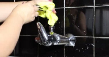 Scrubbing the kitchen faucet with a yellow rag and detergent to clean it, a mature adult cleaning woman reflected in the black tiles, female hands. Cleaning and housework concept