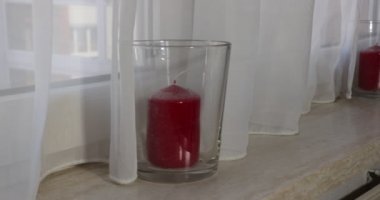Close-up of female hand passing and touching the surface of a very dusty window sill, decorative red candle in a glass candle holder, a transparent white curtain. Cleaning and housework concept