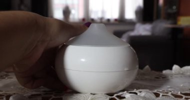 Female hands turning on a white scented aroma oil diffuser humidifier inside a home, against a blurred background, with a color changing lamp. Concept of ambient relaxation at home