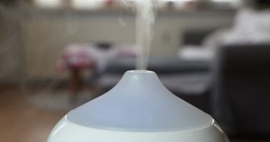 Close-up of the top of a white aroma oil diffuser humidifier exhaling fragrance mist in a home interior, against a blurred background, with a color changing lamp. Concept of ambient relaxation at home