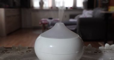 Close-up of a white aroma oil diffuser humidifier exhaling fragrance mist in a home interior, against a blurred background, with a color changing lamp. Concept of ambient relaxation at home