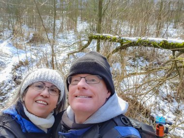 Couple taking a selfie against leafless vegetation and snow on ground, looking up, smiling, wide smile, glasses, winter clothes, crochet headband, black hat, blue jackets, wintry day in Netherlands