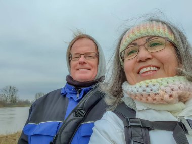 Couple taking a selfie against gray cloudy sky, river in background, looking at camera, smiling, wide smile, glasses, winter clothes, crochet headband and neck, blue jackets, wintry day, Netherlands