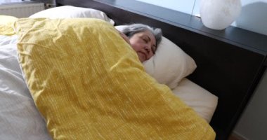 Olgun bir kadın huzur içinde ve yavaşça uyanıyor, yüzüne dokunuyor, pencereden içeri güneş ışığı giriyor, yavaşça hareket ediyor, gri siyah uzun saçlar, sarı yorgan, yeni bir güne başlamak için gülümsüyor.