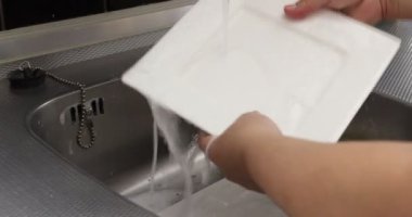 Yetişkin kadın elleri kare beyazı bulaşıkları yıkıyor, akan suda deterjan kaldırıyor, ev mutfağında metal lavabo, sarı sünger. Ev işi kavramı