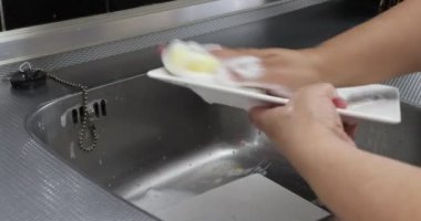 Yetişkin kadın elleri sarı bir süngerle kare beyaz tabakları ovalıyor, deterjan ve su kullanıyor, ev mutfağında metal lavabo kullanıyor. Ev işi kavramı