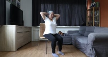 Yaşlı bir kadın belini çalıştırıyor sandalyeye oturuyor, elleri başının üstünde oturuyor ve arka planda bir yandan diğer yana dönüyor. Evdeki olgun insanlar için egzersiz kavramı ve sağlıklı yaşam tarzı