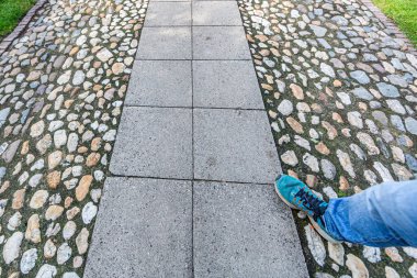 Ortasında kocaman gri tuğlalar olan taşlardan yapılmış bir yol, mavi tenis ayakkabılı bir erkek bacağı kaldırımda yürüyor, yeşil çimenler, bir evin bahçesi, bahar güneşli bir gün.