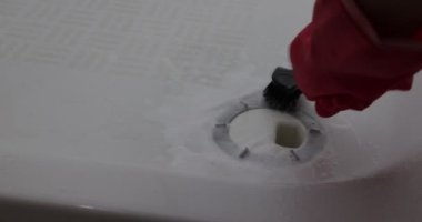Klorlu deterjanla siyah fırçayla ovalanan kırmızı eldivenli eller yıkama sürecinde kirli duş gideri, beyaz metal yüzey, bakım görevi. Temizlik ve ev işi kavramı