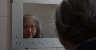 Yaşlı ev hanımı banyo aynasını temizliyor, sıvı temizleyiciyi spreyliyor ve kağıtla ovalıyor, aynanın karşısında dikiliyor, iş memnuniyetini ifade ediyor. Ev temizliği kavramı