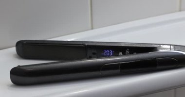 Banyodaki beyaz bir raftaki siyah düzleştirici demire yakın çekim, 201 ile 210 derece arasındaki sıcaklığı, modern yüksek teknoloji aletleri kavramını gösterir.