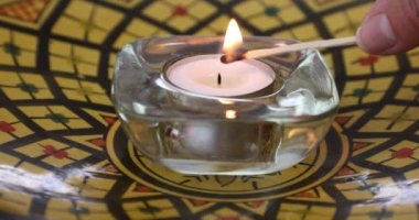 Yetişkin kadın elleri, arka planda siyah kare süslemeli sarı bir tabakta cam bir mum tutacağıyla çay lambasını yakarak huzur hissi veriyor. İnanç ve umut kavramı