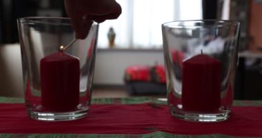Yetişkin kadın elleri cam mum tutacaklarında iki kırmızı mum yakıyor, oturma odasında huzur ve sükunet hissi veriyor, arka planda bulanık bir pencere. İnanç ve umut kavramı