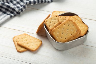 Beyaz ahşap masa arkasında masa örtüsü olan paslanmaz çelik bir kasede kraker kurabiyeleri..