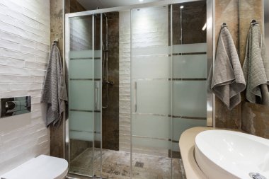 Banyo modern tarzda döşeli döşemeleri ve duvarları var. Cam kapılı iki duş, beyaz tuvalet ve havlu..