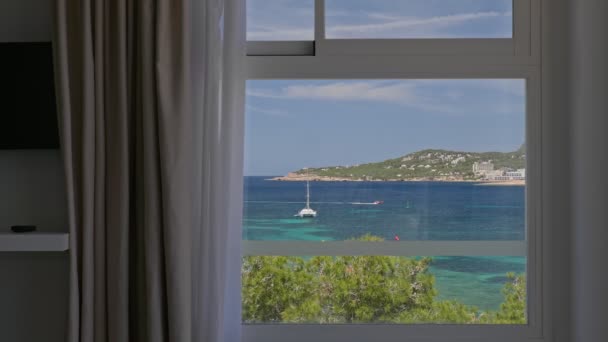 Blick aus dem Fenster eines Hotelzimmers mit Meer, Bäumen, Bergen, Yachten und einem Boot, das auf türkisfarbenem Wasser schwimmt. Ibiza, Spanien. — Stockvideo