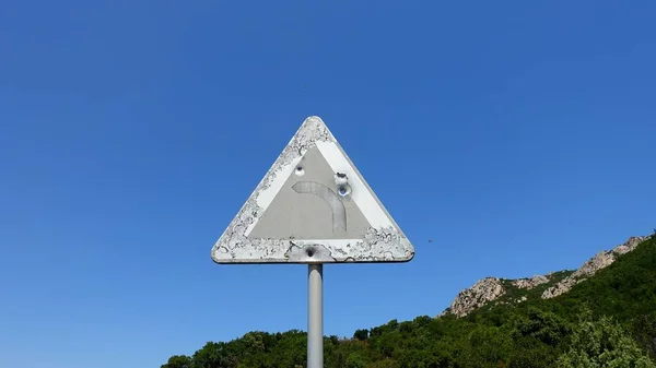 An old road sign on a mountain road indicating a dangerous curve, perhaps riddled with rifle shots.
