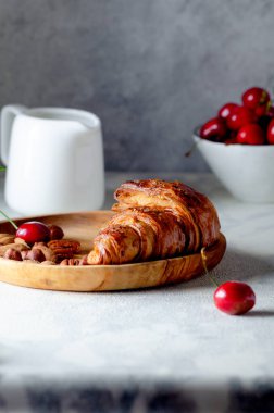 Tahta tabakta taze hamur kruvasanı ve gri arka planda olgun kirazlar..