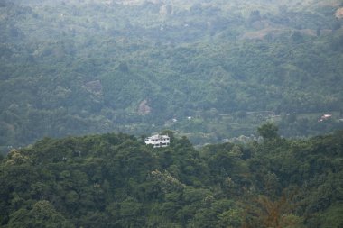 Single house on a big hill drone photography. Mountain landscape aerial view with green trees. Rural hilly landscape photo with a resort building on the top. Beautiful hilltop view with green trees.