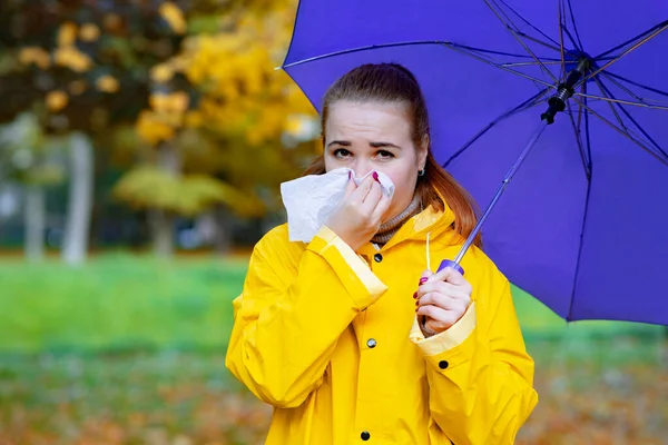 Sick woman blows her nose and sneezes outdoors. During an epidemic of flu and colds, the female suffers from bronchitis or allergies. Girl in autumn with an umbrella and a raincoat — Stock Photo, Image